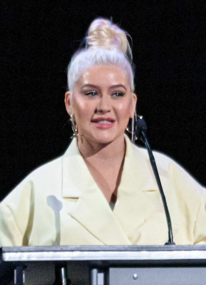 Christina Aguilera speaking at a podium, dressed in light-colored attire with hair styled in a high bun.