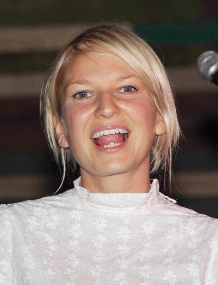 Sia smiling and speaking near a microphone, highlighting key moments from her bio and career highlights.