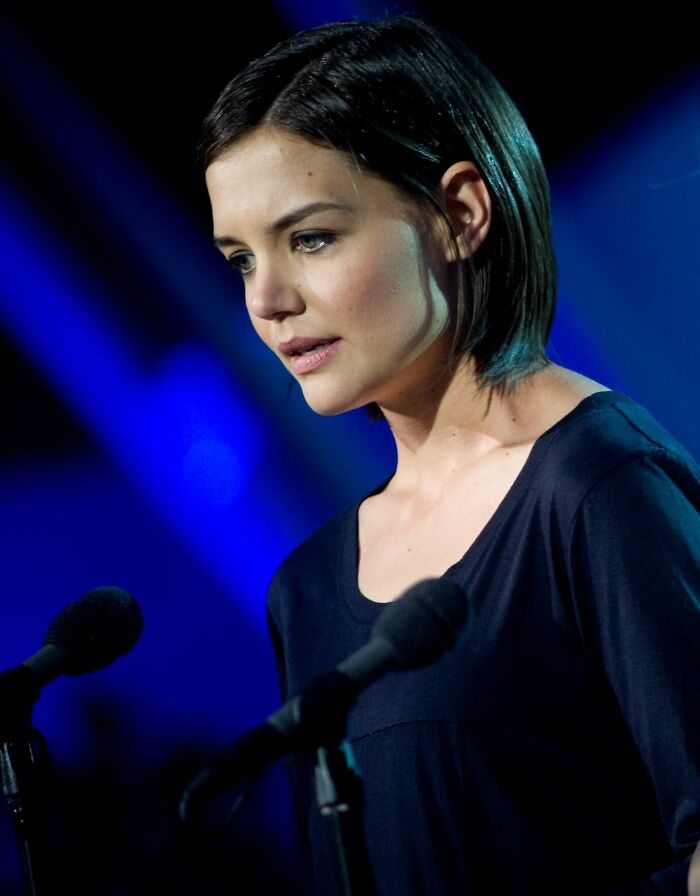 Katie Holmes speaking at an event, dressed in a black top with a focused expression and dark background lighting.