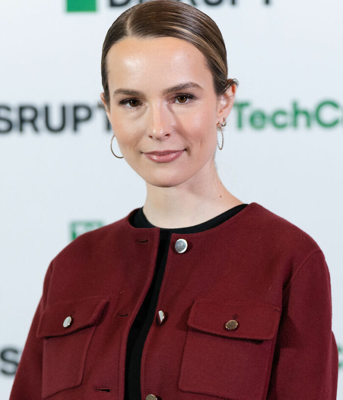 Bridgit Mendler wearing hoop earrings and a burgundy jacket, attending a TechCrunch event with a confident expression.