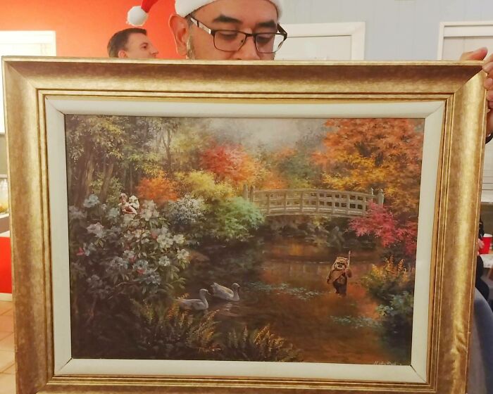 Man wearing Santa hat holding a framed painting with a small Ewok figure by a pond in bizarre Christmas gifts collection