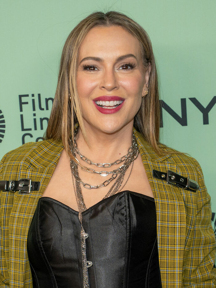 Alyssa Milano smiling at a film event, wearing layered chains, a black top, and a yellow plaid jacket.