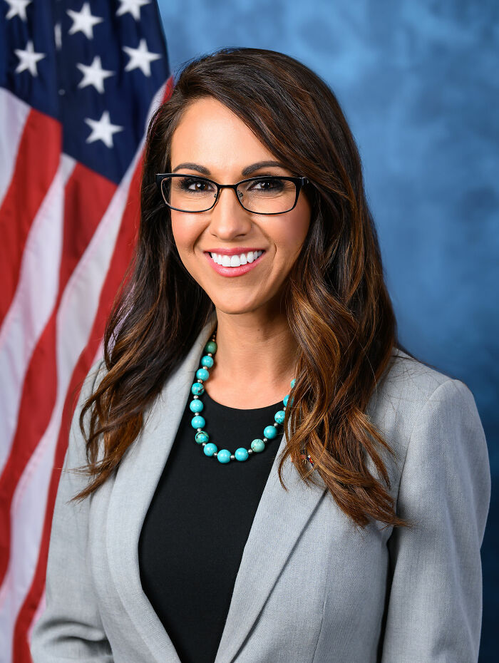 Lauren Boebert smiling, wearing glasses and a turquoise necklace, posed in front of an American flag and blue backdrop.