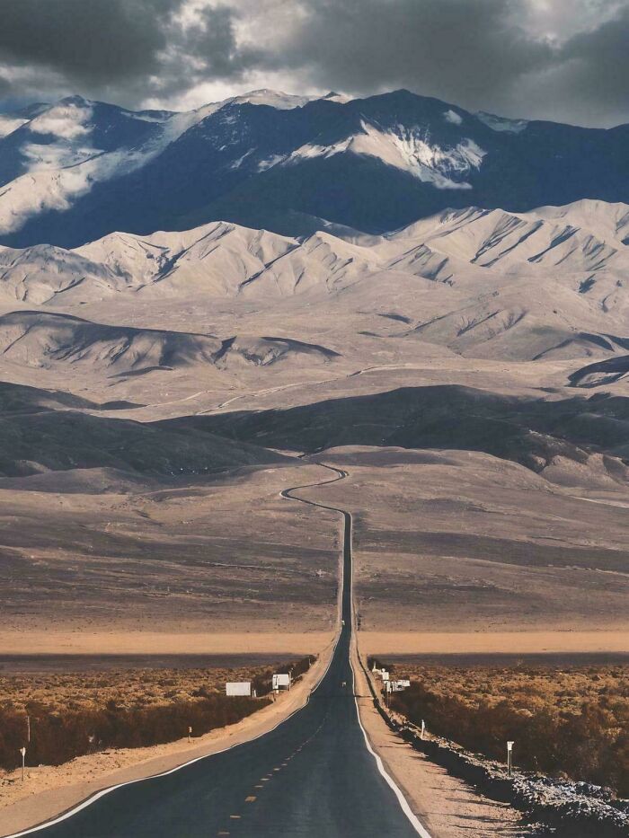Desert road stretching into barren liminal spaces with distant mountains under moody cloudy sky resembling the backrooms.