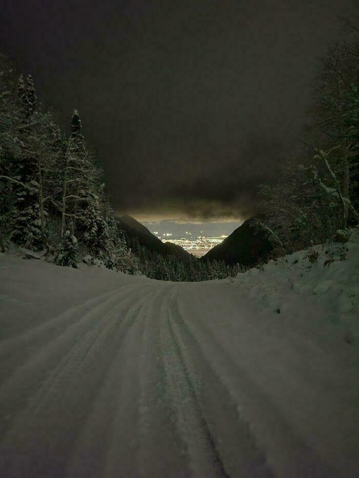 Snow-covered road through dark forested liminal spaces with distant city lights under an ominous cloudy night sky.