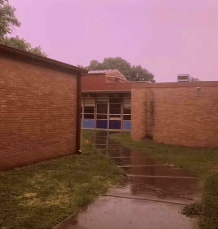 Empty narrow corridor between brick walls with wet pavement and overcast sky, evoking liminal spaces like the Backrooms.