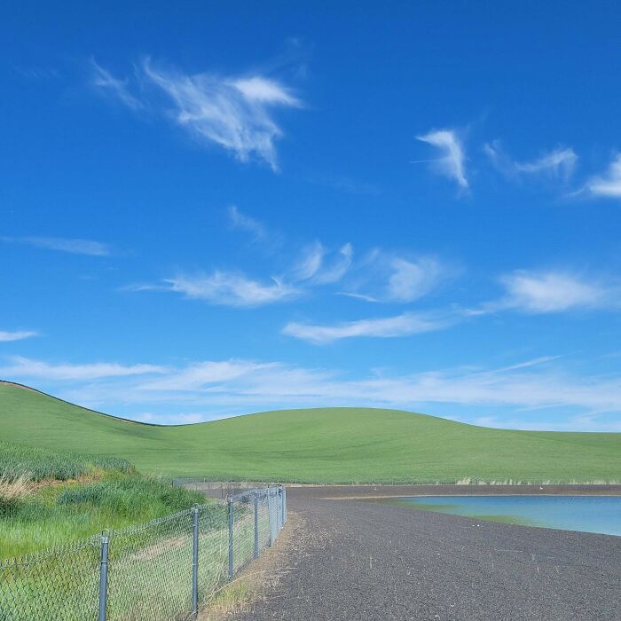 Calm liminal space with a green hill, clear blue sky, and a fenced path by the water, resembling the backrooms atmosphere.