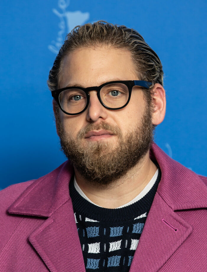 Jonah Hill with glasses and beard, wearing a patterned sweater and purple coat against a blue background.