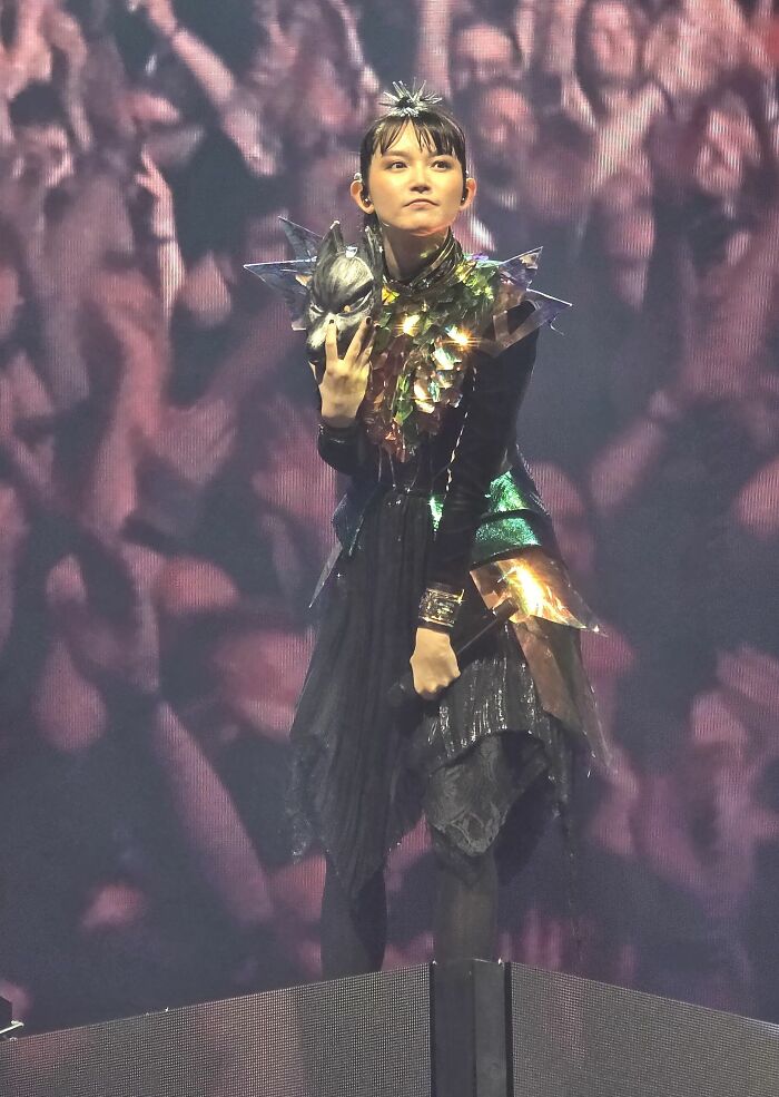 Suzuka Nakamoto performing on stage in a futuristic outfit holding a mask during a live concert event.
