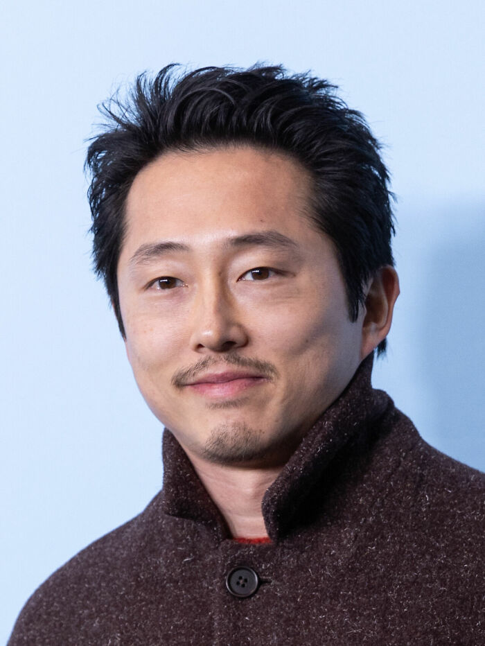 Steven Yeun smiling, wearing a dark coat, with a light blue background, representing bio and career highlights.