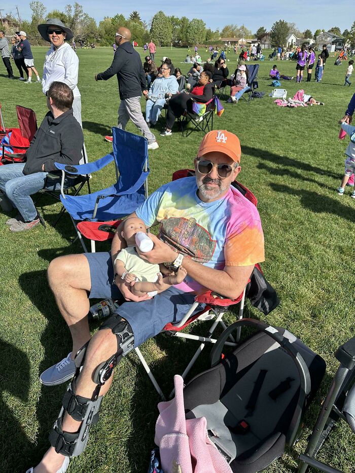 Man with leg brace feeding baby from bottle, wearing colorful shirt and orange cap at outdoor gathering, funny pictures from 2025.