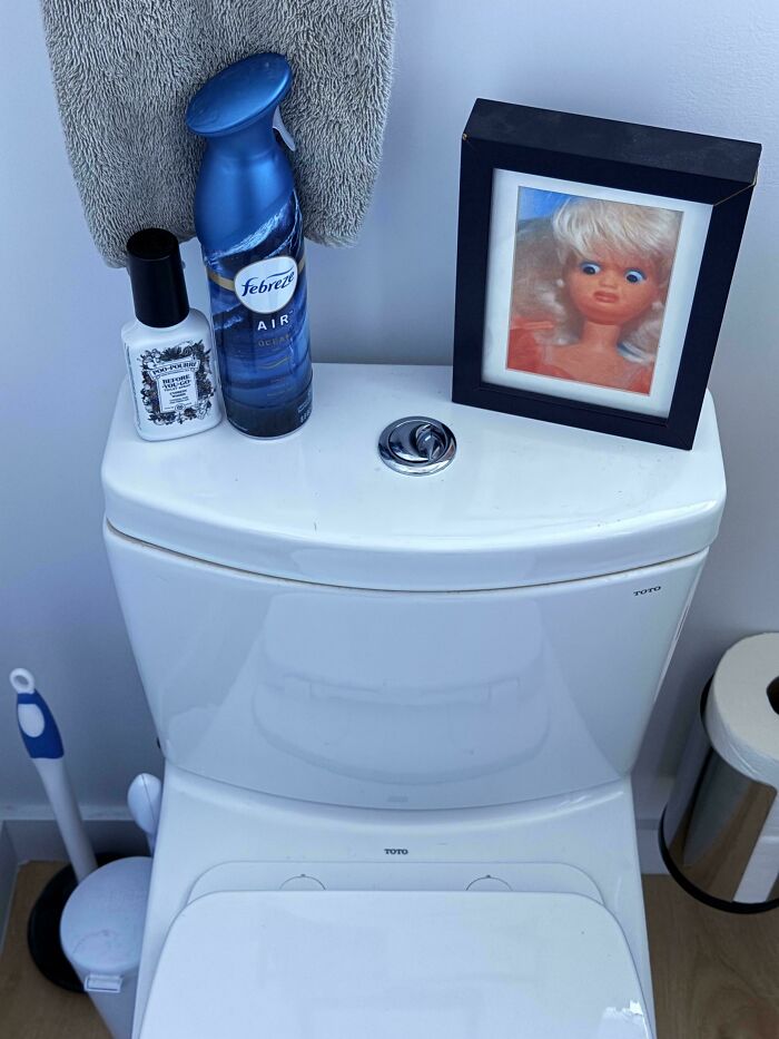 Bathroom toilet tank with air freshener, hand lotion, framed funny doll picture, brush, and toilet paper holder nearby.