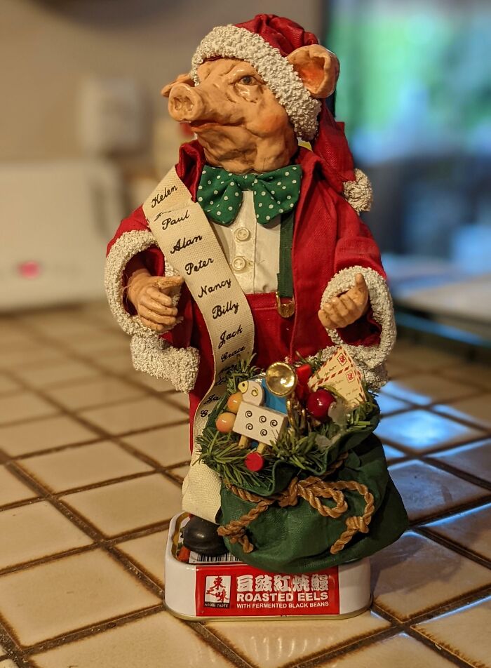 Pig figurine dressed as Santa holding a list and a bag on a roasted eels tin, bizarre Christmas gift idea.