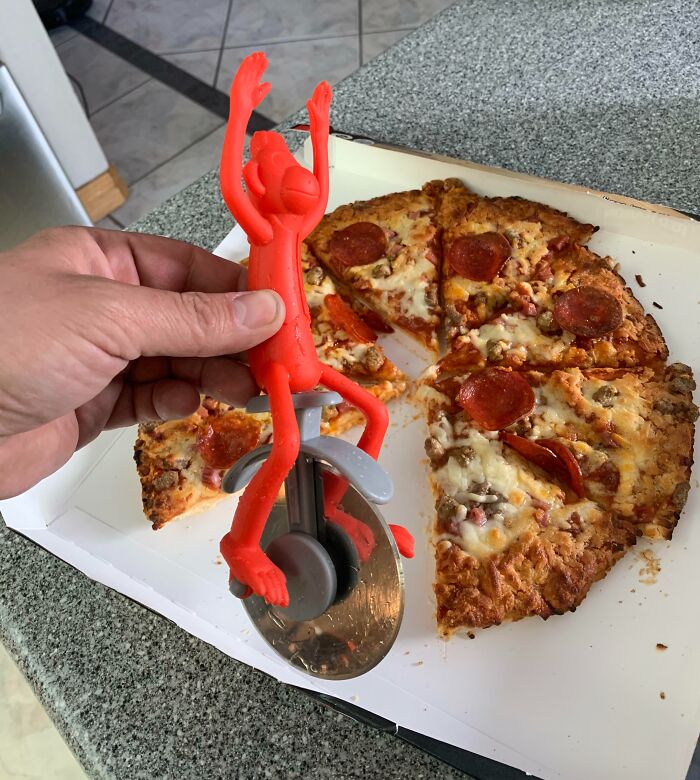 Hand holding a bizarre red monkey pizza cutter over a partially eaten pepperoni pizza on a kitchen counter.