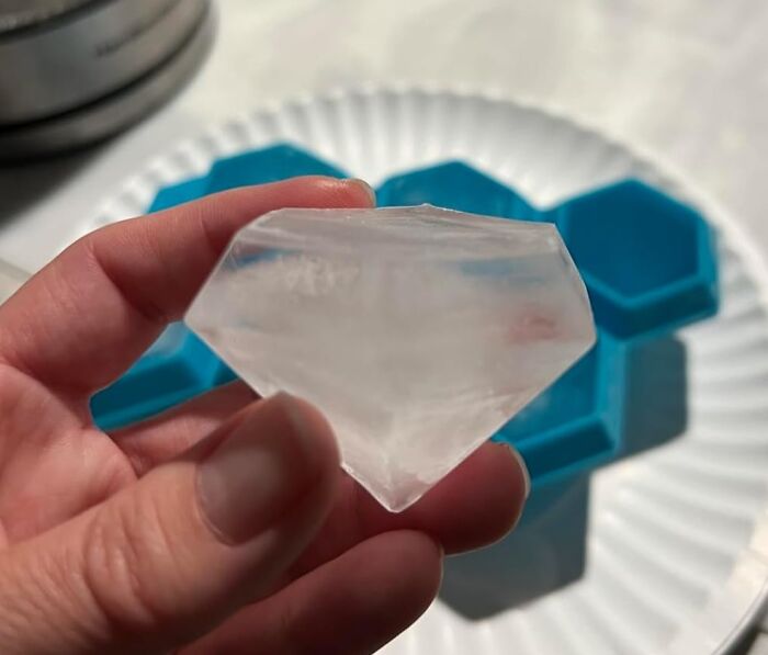 Hand holding a clear diamond-shaped ice cube beside a blue hexagonal silicone mold on a white plate, best sellers 2025.
