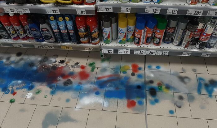 Store spray-paint aisle with cans on shelves and colorful paint splattered across tile floor, next-level jerks vandalism