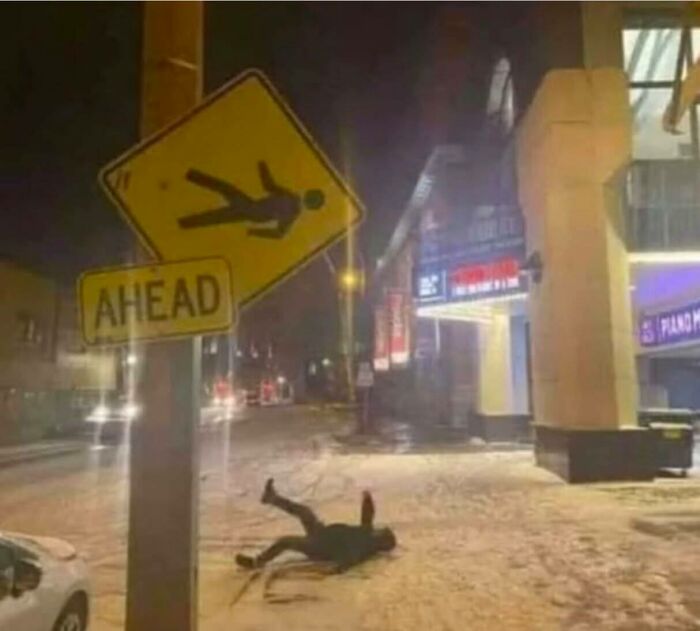 Person lying on snowy street under a funny stupid signs pic showing a falling figure ahead warning at night.