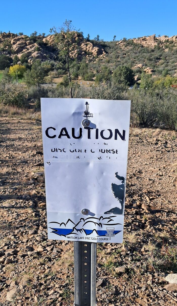 Worn and partially torn caution sign at a disc golf course in a rocky, natural outdoor setting showing funny stupid signs pics.