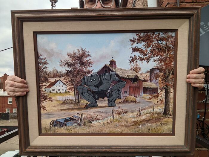 Person holding thrifted painting of a rustic farm with a funny and unexpected cartoon robot added inside.