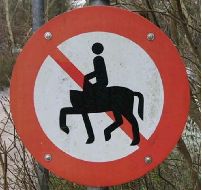 No horse riding allowed sign with a funny-stupid design showing a person riding a four-legged animal in a wooded area.