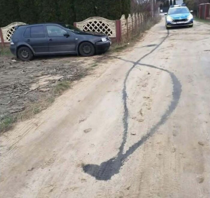 Tire skid marks on a dirt road creating a humorous shape in a rural Eastern Europe setting with parked cars nearby.