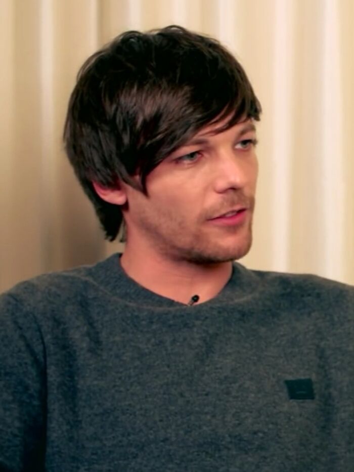 Louis Tomlinson in a casual grey sweater, engaged in conversation during an indoor interview setting.
