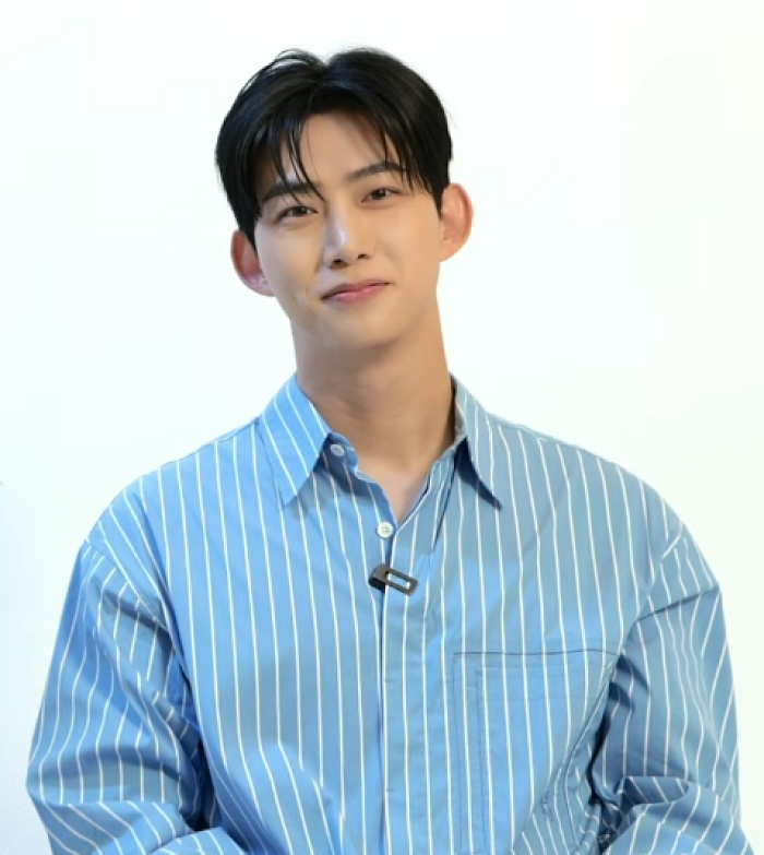 Ok Taec-yeon wearing a blue striped shirt with a microphone, smiling gently against a plain white background.
