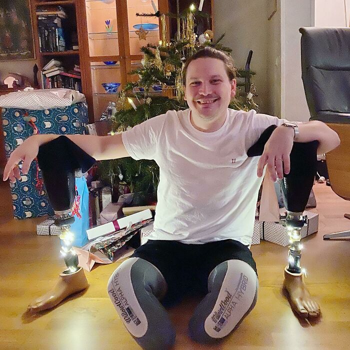 Young man with prosthetic legs sitting happily on the floor by a decorated Christmas tree in a heartwarming holiday moment.