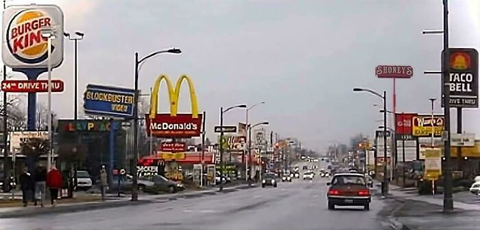 A busy street lined with 90s and 2000s nostalgia fast food and retail signs like McDonald's and Blockbuster.
