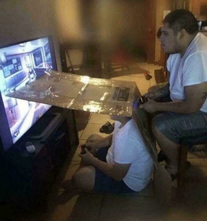 Two people playing video games with a cardboard shield between them, evoking 90s and 2000s nostalgia.