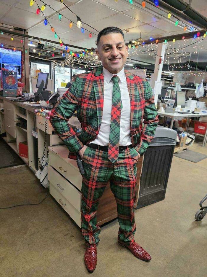 Man in a colorful holiday plaid suit and tie smiling in a festively decorated indoor setting with string lights.