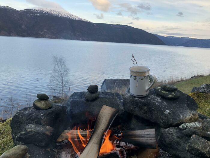 Cozy holiday moment by a lakeside campfire with a mug of hot drink and snowy mountains in the background.