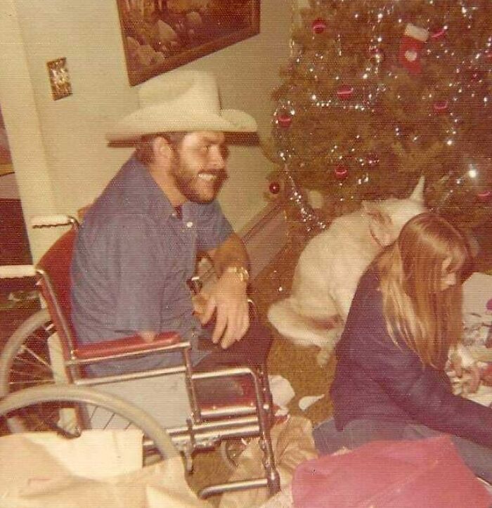 Man in wheelchair wearing cowboy hat smiling near Christmas tree with child and dog during holiday moments so sweet.
