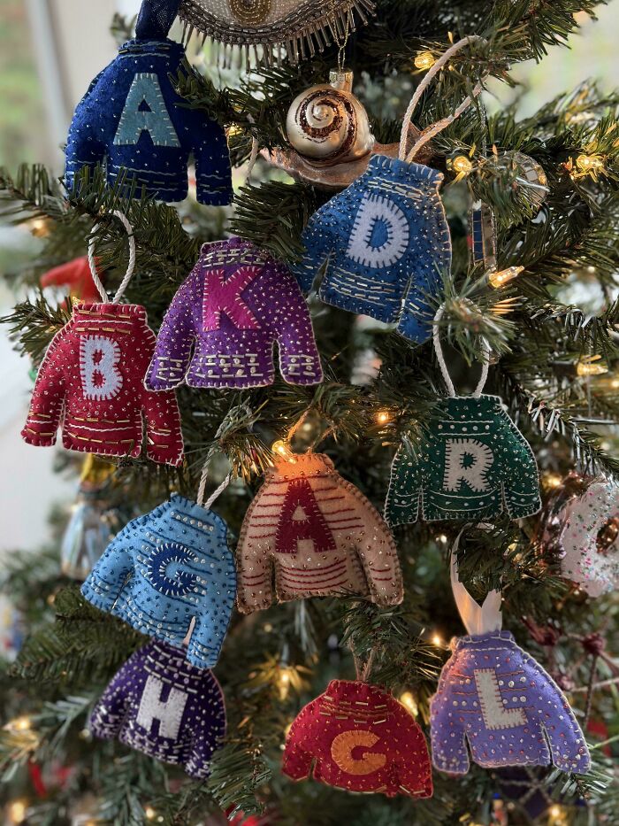 Colorful handmade felt holiday sweater ornaments with letters hanging on a decorated Christmas tree with lights.