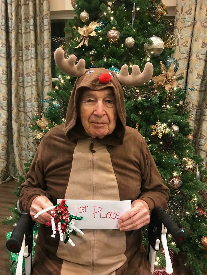 Elderly man in a reindeer costume holding a 1st place sign in front of a decorated Christmas tree holiday moments.