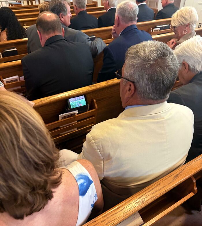 Man in a white jacket watching a football game on phone during a formal event, funny pictures capturing the weirdness of 2025.