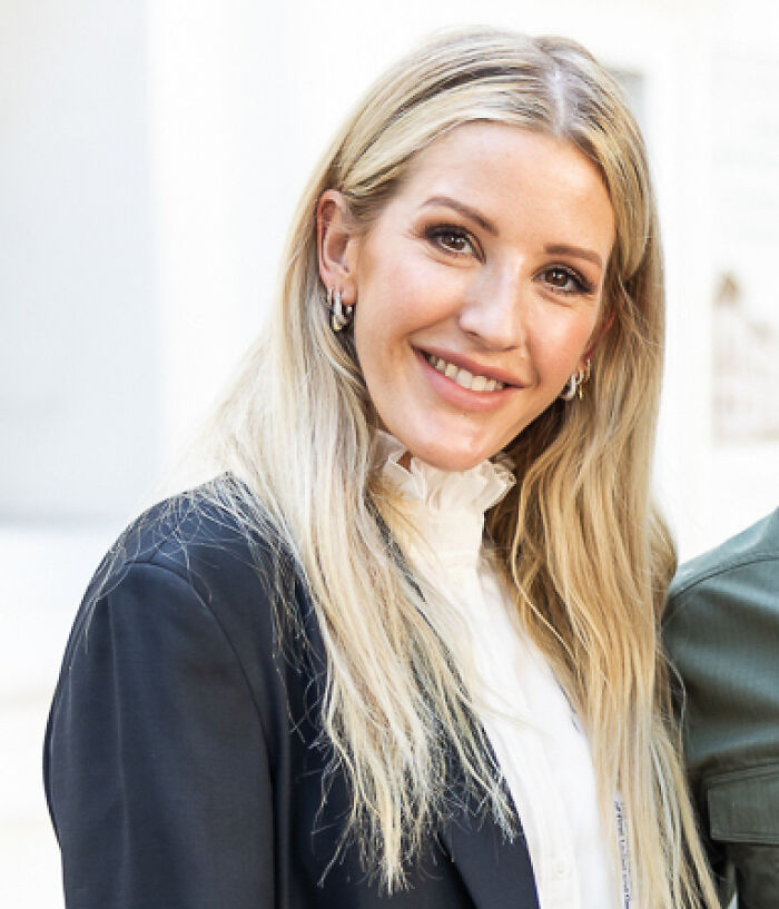 Ellie Goulding smiling, wearing a black blazer and white blouse, highlighting her bio and career achievements.