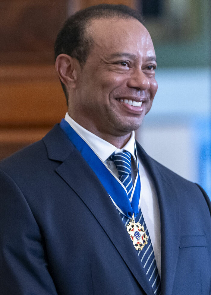 Tiger Woods smiling in a suit wearing a medal, highlighting achievements in his bio and career highlights.