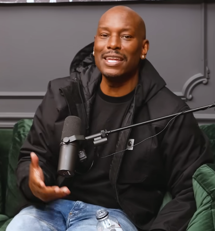 Tyrese Gibson speaking into a microphone during a podcast, wearing a black jacket and jeans in a studio setting