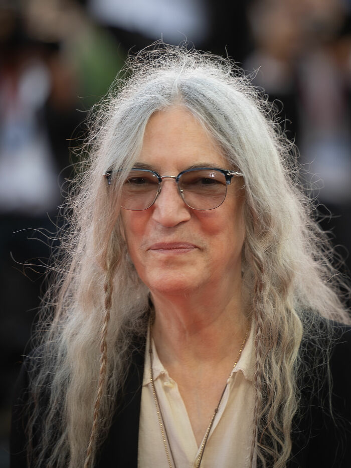 Patti Smith with long gray hair and glasses, smiling at a public event with blurred background.
