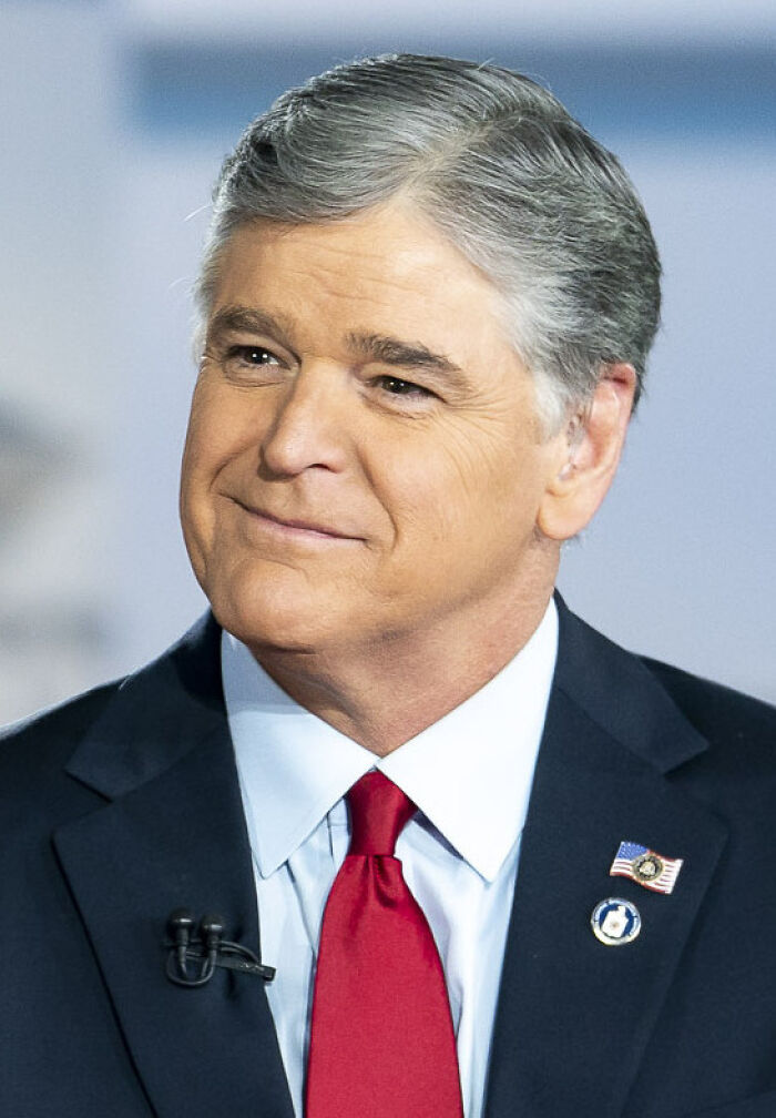 Sean Hannity in a dark suit and red tie with US flag pin, appearing in a professional broadcast studio setting.