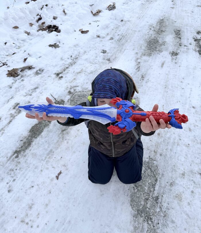 Child kneeling on snowy ground holding a large colorful toy sword, capturing funny pictures from 2025 weird moments.