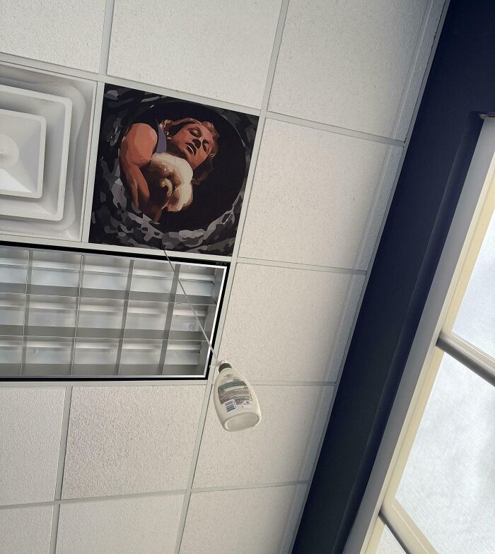 Ceiling tile replaced with art of person holding a cat, with a hanging bottle of lotion in a funny picture from 2025 capturing weirdness.