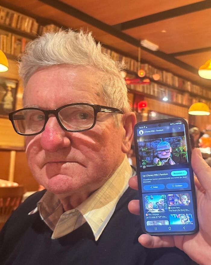 Elderly man with glasses sitting in a cozy restaurant, someone holding a phone showing a funny picture from 2025.