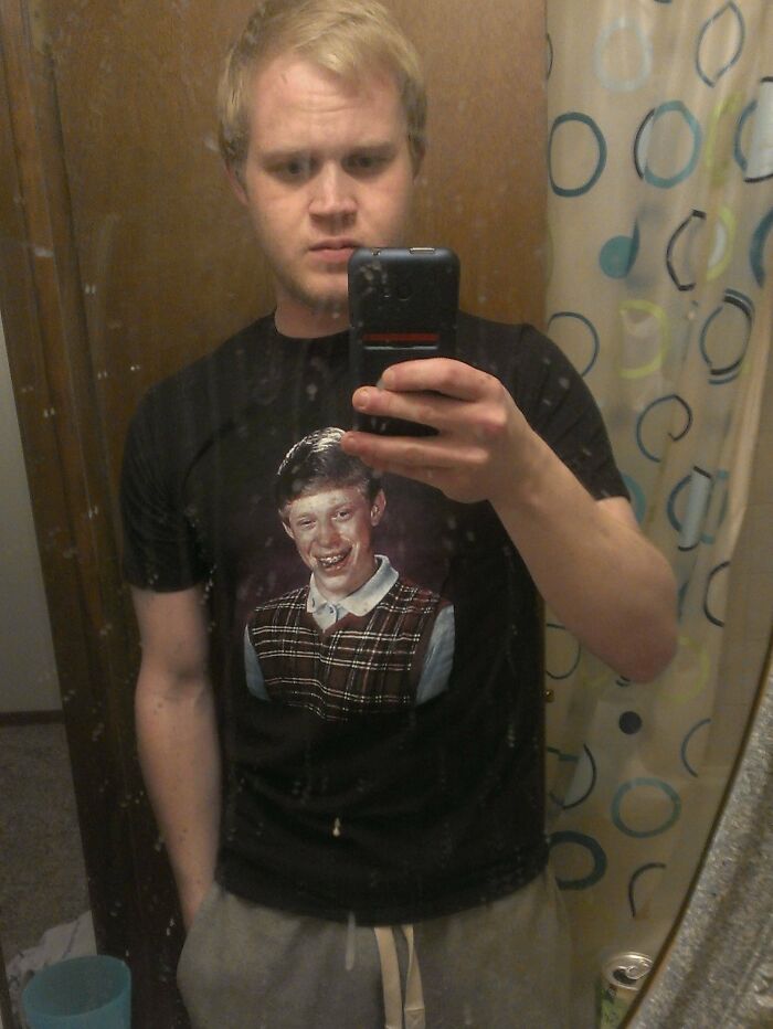 Man taking mirror selfie wearing a hilariously bad Secret Santa gift t-shirt featuring an awkward school photo.