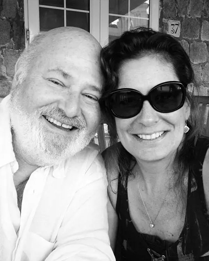Rob Reiner smiling in a casual setting posing with a woman wearing dark sunglasses outdoors by a stone wall.