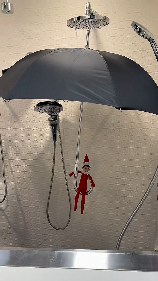 Elf on the Shelf hanging from a showerhead holding an umbrella in a bathroom for playful mischief.