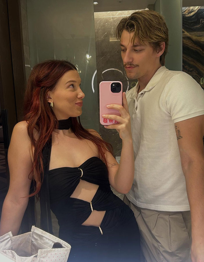 Millie Bobby Brown taking a mirror selfie with a man, showing her arm injury that fuels fan theories about Jake Bongiovi control.