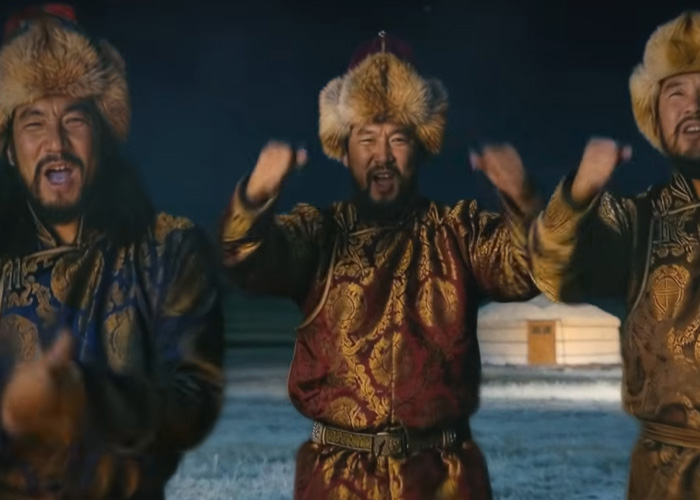 Three men in traditional Mongolian attire and fur hats performing throat singing outdoors at night near a yurt in winter.