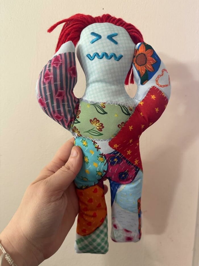 Hand holding a patchwork doll with red yarn hair, illustrating gag gifts for the friend who thinks a scented candle is a cry for help.